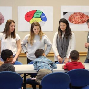 Four students stand around a table with children. Brain posters hang on the wall behind them.